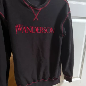 Svart JW Anderson sweatshirt stl S (knappt använd) - Svart sweatshirt från JW Anderson i 100% bomull med röd broderad logga framtill och röda kontrastsömmar. Klassisk rund halsringning och långa ärmar. Perfekt för dig som gillar stilrena men ändå unika detaljer. Skön streetwear-stil som ändå är dressad. Tröjans design är att vara ”ut och in”. 