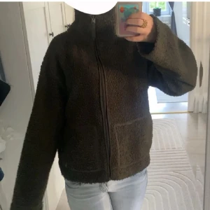 Mörkgrön teddyjacka med dragkedja - Mysig teddyjacka med hög krage och hel dragkedja framtill. Jackan har två stora fickor och en loose passform som ger en avslappnad vibe. Perfekt för kyliga dagar när du vill vara både varm och snygg.