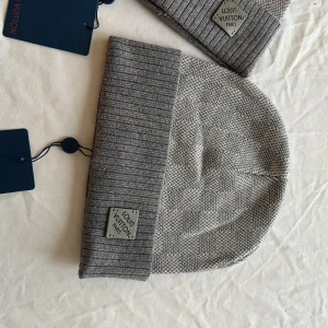 Grå rutig Louis Vuitton mössa - Exklusiv grå mössa  Louis Vuitton med klassiskt rutigt mönster och bred ribbad kant. Mössan har en metallbricka med Louis Vuitton Paris på framsidan och LV-logga på etiketten. Materialet är mjuk ull som håller dig varm och stilsäker.
