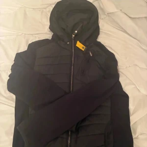 Svart pufferjacka från Parajumpers - Svart pufferjacka från Parajumpers med huva och dragkedja framtill. Jackan har quiltade partier, en gul detalj vid halsen och orange detaljer på insidan. Perfekt för kalla dagar och har en clean, sportig look.