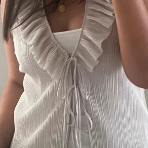 Plisserad volang blus - Säljer en vit blus med ribbad struktur, djup v-ringning och bred volangkant runt halsen. Toppen har snörning framtill och är ärmlös, vilket ger en chill och feminin vibe. Perfekt för dig som gillar detaljer och vill sticka ut lite extra.