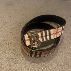 Burberry bälte med rutigt mönster - Säljer ett snyggt Burberry bälte med klassiskt rutigt mönster i beige, brunt och svart. Bältet har en stilren metallspänne med Burberrys ikoniska 'B'-logga. Utsidan är i svart skinn och insidan har det typiska Burberry-rutiga mönstret. Perfekt för att lyfta din outfit.