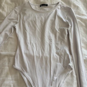 Vit långärmad bodysuit från Zara - Säljer en stilren vit bodysuit från Zara i storlek S. Modellen har rund halsringning och långa ärmar, perfekt för att skapa en clean och enkel look. Materialet är mjukt och stretchigt, vilket gör den bekväm att bära hela dagen.
