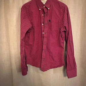 Rutig röd skjorta Abercrombie & Fitch - Snygg rutig skjorta från Abercrombie & Fitch i rött och mörkblått. Klassisk button-down krage, långärmad och med knappar framtill. Skjortan har en bröstficka med broderad logga och är gjord i mjuk bomull. Perfekt för dig som gillar en chill och stilren look.