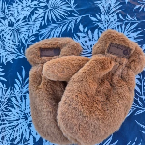 Bruna pälsvantar A Baby Bear - Supermjuka och fluffiga bruna vantar från A Baby Bear. Vantarna är täckta av fejkpäls och har en söt etikett med en björn på mudden. Perfekta för kalla dagar när du vill hålla händerna varma och se gullig ut.