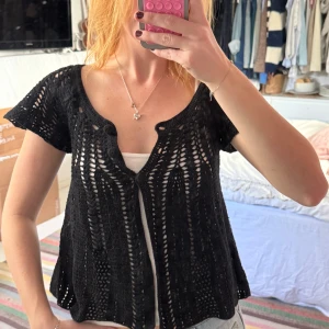 Svart virkad blus med knappar - Säljer en svart virkad blus med korta ärmar och snyggt mönster. Blusen har tre knappar framtill och är lätt transparent, perfekt att ha över ett linne. Den har en luftig och avslappnad passform med fina virkade detaljer.