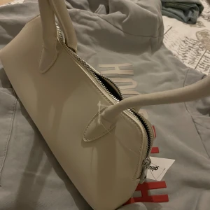 Vit handväska från Gina Tricot - Stilren vit handväska från Gina Tricot med mjukt syntetmaterial och silverfärgad dragkedja. Väskan har två rejäla handtag och en minimalistisk design som passar till många olika outfits. Köpt för 299 , pris kan diskuteras 