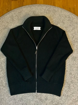 Svart stickad kofta från Maison Margiela - Svart ribbstickad kofta från Maison Margiela med hög krage och dubbel dragkedja framtill. Ikoniska vita stygn i nacken. Grovstickad känsla och långärmad modell, perfekt för lager-på-lager. Snygg och stilren design som funkar till allt.