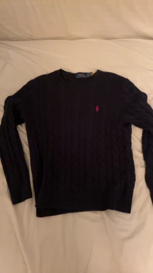 Mörkblå stickad tröja från Polo Ralph Lauren - Säljer en mörkblå stickad tröja från Polo Ralph Lauren med klassiskt kabelstickat mönster och röd broderad logga på bröstet. Tröjan har rund hals och långa ärmar, perfekt för dig som gillar preppy stil. Helt utan defekter.