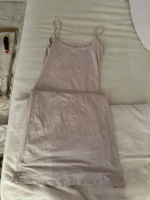 Ljusbeige långklänning med smala band - En enkel och stilren långklänning i ljusbeige ton med smala axelband och rak passform. Klänningen är ärmlös och har en mjuk, slät yta som känns skön mot huden. Perfekt för varma dagar eller som ett lager under andra plagg.