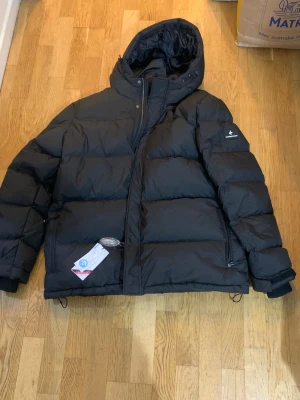 Svart pufferjacka Cross Sportswear XXL - Svart pufferjacka från Cross Sportswear i storlek XXL. Jackan har huva, dragkedja och knappar framtill, samt ribbade muddar vid ärmslut. Fylld med dun och fjäder, yttermaterial i polyester. Perfekt för kalla dagar med sin tjocka och fluffiga look.