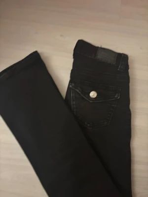 Svarta jeans från Gina Tricot, stl 32 - Svarta utsvängda jeans från Gina Tricot, snygga detaljer som silverfärgade knappar och ficklock bak. Jeansen är tillverkade i ett mjukt denimtyg som sitter skönt och passar till det mesta.