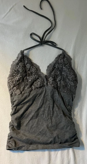 Grå spetsdetaljerad linnetopp - Säljer en grå linnetopp med tunna knytband i nacken och v-ringad spets upptill. Toppen har blommönstrad spets över bysten och är tillverkad i mjukt bomullsmaterial. Perfekt för en trendig och avslappnad look. Storlek xs-S (kanske m) 