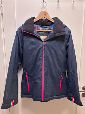 Skidjacka i jeansfärg från Helly Hansen - Snygg mörkblå täckjacka från Helly Hansen med rosa detaljer och dragkedjor. Jackan har huva, Helly Tech Performance-material som är vattentätt och andas, samt RECCO-reflektor. Insidan har färgglatt rutigt foder. Perfekt för dig som gillar att vara ute även när det blåser och vill synas i skidbacken.