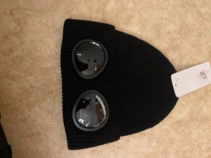 Svart mössa med goggles från C.P. Company - Säljer en svart ribbstickad mössa från C.P. Company med två coola svarta goggles framtill. Mössan har en klassisk form och är tillverkad i stickad material som ger en skön passform. Perfekt för dig som vill sticka ut med streetwear-vibe.