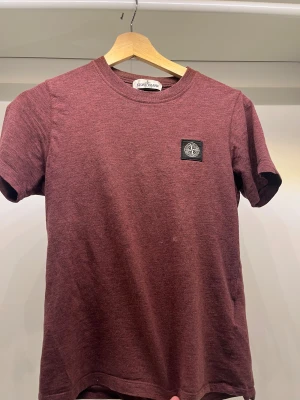 Vinröd t-shirt från Stone Island - Snygg vinröd t-shirt från Stone Island med klassisk rund hals och korta ärmar. T-shirten har en svart patch med Stone Island-loggan på bröstet och är gjord i mjuk bomull. Perfekt för dig som gillar streetwear och vill ha något stilrent med cool detalj.