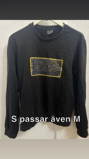 Svart långärmad tröja EA7 Armani - Svart långärmad tröja från EA7 Emporio Armani med stort tryck i gult och grått framtill. Klassisk rund halsringning och mjukt bomullsmaterial. Perfekt för dig som gillar stilrena och sportiga plagg med tydlig logga.