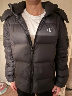 Svart pufferjacka från Calvin Klein - Svart pufferjacka från Calvin Klein med stor huva och logga på bröstet och ärmen. Jackan har dragkedja framtill och två sidofickor med dragkedja. Den är quiltad och har en loose passform, perfekt för kalla dagar. Inga märken eller skador på den alls. Priset går att diskutera vid snabbköp.