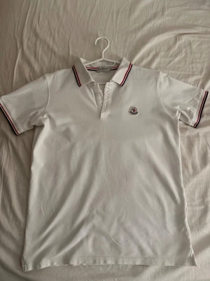 Vit Moncler pikétröja  - Vit pikétröja från Moncler i bomull med klassisk krage och knäppning framtill. Röd och marinblå rand på krage och ärmslut samt Moncler-logga på bröstet. Perfekt för en clean och sportig look.