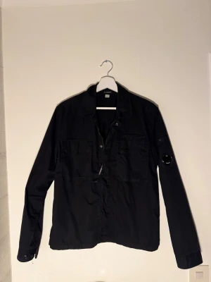 Svart overshirt från C.P. Company - Svart overshirt från C.P. Company med klassisk krage, två stora bröstfickor och ikonisk linsdetalj på vänster ärm. Jackan har både dragkedja och knappar framtill, vilket ger en cool och funktionell look. Perfekt för lager-på-lager streetwear. Dock saknas en knapp på vänster arm men den är sparad!🙂