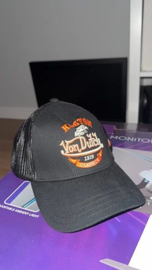 Svart Von Dutch keps  - Säljer en Von Dutch med broderad logga i orange och vitt framtill. Kepsen har mesh på sidorna och bak, samt justerbar snapback. Märket syns även på insidan med tryckta band. 