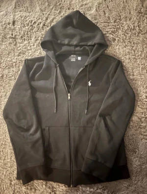 Svart hoodie från Polo Ralph Lauren - Svart hoodie från Polo Ralph Lauren med dragkedja och huva. Tröjan har snörning vid huvan, två fickor framtill och den klassiska vita Polo-loggan broderad på bröstet. Tillverkad i mjuk bomull för skön komfort och stilren look.