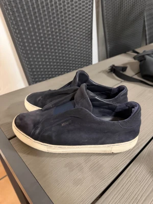 Mörkblå sneakers från Arigato - Snygga mörkblå sneakers från Arigato med stilren design och vit platt sula. Skorna är tillverkade i mocka och har en elastisk detalj över vristen för extra komfort. Perfekta för dig som gillar minimalistisk stil och vill ha något som sticker ut lite. Sulorna har tyvärr hål 