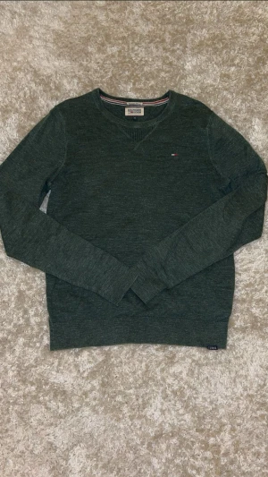 Mörkgrön tröja från Hilfiger Denim - Snygg mörkgrön tröja från Hilfiger Denim med diskret logga på bröstet. Tröjan har v-ringad hals och ribbade muddar vid ärmslut och nederkant. Materialet är mjukt och känns som bomull, perfekt för dig som gillar stilrena och klassiska plagg. 