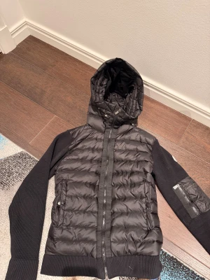 Svart Moncler pufferjacka med huva - Svart pufferjacka från Moncler med stickade ärmar och huva. Jackan har dragkedja framtill, ribbade muddar och fickor med både dragkedja och knapp. Moncler-logga på ärmen och snygga detaljer i metall. Plasten på dragkedjorna är fortfarande på, oanvänd!