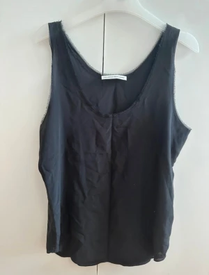 Svart linnetopp från FWSS - Svart linnetopp från FWSS Fall Winter Summer Spring med råa kanter och rundad halsringning. Toppen är ärmlös och har en enkel, loose passform. Materialet känns lätt och svalt, perfekt för varma dagar. Snygg att styla med jeans eller kjol för en chill look