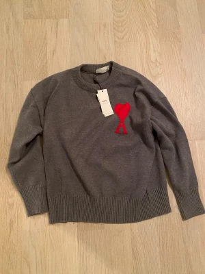 Grå stickad tröja från Ami Paris - Snygg grå stickad tröja från Ami Paris med rund hals och ribbade muddar. På bröstet finns ett stort rött hjärta med bokstaven A, vilket ger en unik och trendig look. Perfekt för dig som gillar stilrena detaljer och vill sticka ut.