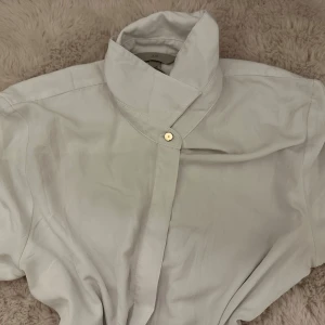 Vit skjorta med hög krage från H&M - Vit skjorta från H&M med unik hög krage och dold knäppning framtill. Skjortan har långa ärmar med manschett och en stilren, minimalistisk look. Perfekt för dig som gillar clean och modern stil.