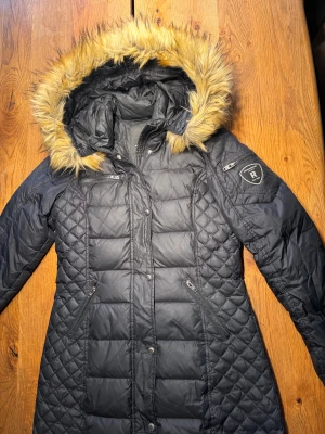 Svart dunjacka Rockandblue med fuskpäls - Svart dunjacka från Rockandblue med snygg quiltad design och fuskpäls på huvan. Jackan har flera dragkedjor, fickor och Rockandblue-logga på ärmen. Perfekt för kalla dagar med sin långa passform och värmande material. 