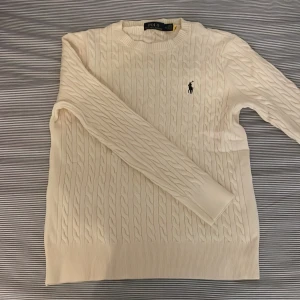 Vit kabelstickad tröja Polo Ralph Lauren - Snygg vit kabelstickad tröja från Polo Ralph Lauren med klassisk rund hals och broderad logga på bröstet. Tröjan har långärmad passform och ribbade muddar vid hals, ärmslut och nederkant. Priset är inte hugget i sten 😁