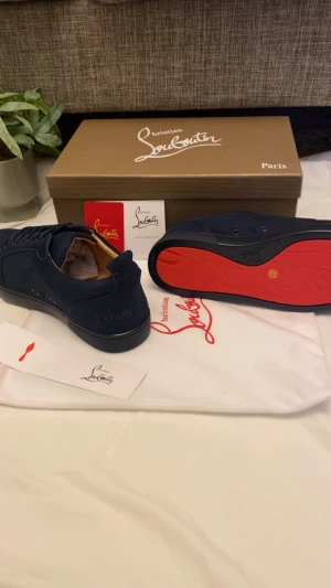 Mörkblå sneakers från Christian Louboutin - Snygga mörkblå sneakers från Christian Louboutin med klassisk röd sula och diskret logga på sidan. Skorna har rund tå, platt sula och snörning. Utsidan är i mocka och insidan i skinn, vilket ger en lyxig känsla. Perfekt för dig som gillar exklusiva detaljer och stilren design.