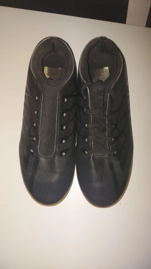 Svarta Umbro fotbollsskor inomhus - Säljer ett par svarta Umbro fotbollsskor för inomhusbruk med gummisula i orange. Skorna har snörning och en stilren design med diskreta Umbro-loggor. Ovandelen är gjord i syntetmaterial med mönstrade detaljer och rund tå. Perfekt för futsal eller träning inomhus. Men på en par så saknas det snöre.