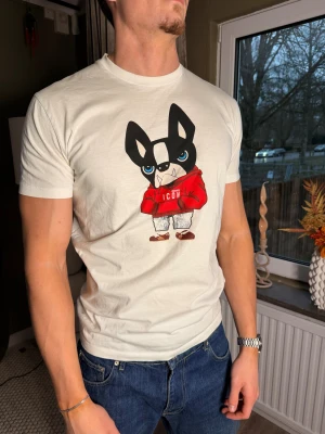 Vit t-shirt med hundtryck från Dsquared2 - Snygg vit t-shirt från Dsquared2 med ett stort tryck av en tecknad hund i röd hoodie på bröstet. Klassisk passform med rund hals och korta ärmar. Tillverkad i 100% bomull för en skön känsla. Perfekt för dig som gillar statement-plagg.