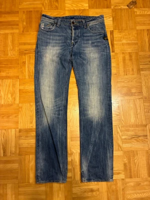 G-Star Raw Attacc Low Straight jeans - Snygga blå jeans från G-Star Raw, modell Attacc Low Straight. Klassisk femficksdesign med vit GS RAW-tryck på myntfickan och slitna detaljer för en cool look. Raka ben och låg midja, tillverkade i robust denim. Perfekt för dig som gillar streetwear.