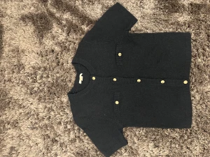 Marinblå kofta med guldfärgade knappar Gina Tricot - Kortärmad marinblå kofta från Gina Tricot med snygga guldfärgade knappar framtill och på de två dekorativa fickorna. Koftan är stickad och har ribbade kanter vid hals, ärmslut och nederkant. Perfekt för dig som gillar klassisk och stilren look.