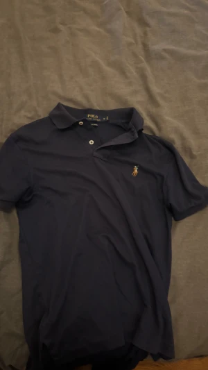 Mörkblå piké från Polo Ralph Lauren - Snygg mörkblå pikétröja från Polo Ralph Lauren i storlek S. Klassisk krage, två knappar framtill och den ikoniska broderade loggan på bröstet. Tillverkad i mjuk bomull som känns skön mot huden. Perfekt för dig som gillar stilren och enkel design.skriv gärna vid frågor. Pris kan diskuteras.