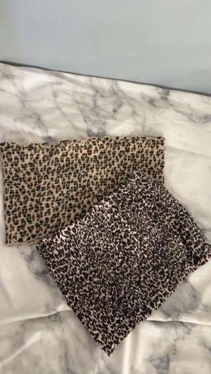 Leopardmönstrade bandeau tubtoppar - Två bandeau tubtoppar med leopardmönster i beige och brun/svart. Båda är smockade och har en elastisk passform som sitter tight mot kroppen. Perfekt för dig som vill ha en edgy och trendig look med djurprint. Säljes tillsammans eller separat.