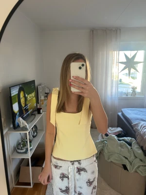 Gul korsett-topp med knytband - Säljer en ljusgul korsett-topp med breda axelband som knyts i rosetter på axlarna. Toppen har en snygg, figurnära passform och fyrkantig ringning både fram och bak. Materialet känns stadigt och har en slät yta. Perfekt för dig som vill ha en trendig och söt look.