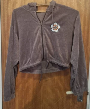 Brun croppad hoodie från Santa Cruz - Mysig brun croppad hoodie från Santa Cruz med dragkedja framtill och snörning i midjan. Tröjan har huva, långa ärmar och ett broderat Santa Cruz-märke på bröstet. Perfekt för en avslappnad streetstyle-look.