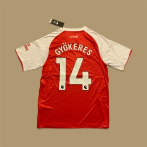 Arsenal Gyökeres #14 matchtröja - Snygg Arsenal fotbollströja med Gyökeres och nummer 14 på ryggen. Röd kropp med vita korta ärmar och vita detaljer, Adidas-logga och klubbmärke på bröstet. Materialet är lätt och ventilerande med subtilt mönster i tyget. Perfekt för dig som älskar Premier League.