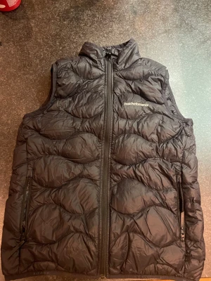 Svart dunväst från Peak Performance - Svart dunväst från Peak Performance med coolt quiltat mönster och hög krage. Västen har två sidofickor med dragkedja och Peak Performance-logga på bröstet. Perfekt att slänga över en hoodie när det är kyligt ute.