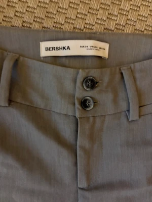 Grå bootcut kostymbyxor från Bershka - Snygga grå kostymbyxor från Bershka med bootcut-snitt och två knappar i midjan. Byxorna har bälteshällor och är tillverkade i ett mjukt, vävt material som ger en clean och stilren look. Perfekt för dig som gillar klassisk stil med modern touch.