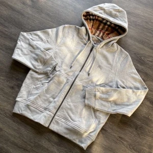 Grå hoodie från Burberry med huva - Säljer en grå hoodie från Burberry med klassiskt rutigt foder i huvan. Tröjan har dragkedja, snörning vid huvan och två fickor framtill. Perfekt för dig som gillar stilrena och bekväma plagg med en touch av lyx.