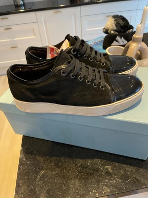 Svarta Lanvin sneakers med lackad tå - Lanvin sneakers i svart mocka och kalvskinn med snyggt lackad tå och vit platt sula. Klassisk rund tå och snörning. Kommer med originalkartong, extra svarta skosnören och dustbag. Perfekt för dig som gillar stilrena och lyxiga sneakers. Skorna är i uk6 priset går att diskutera!