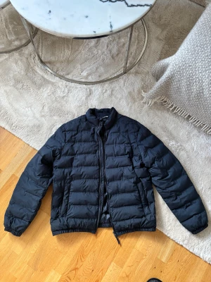 Mörkblå jacka från Zara strl M - Snygg svart pufferjacka med klassisk ståkrage och hel dragkedja framtill. Jackan har quiltad design, två sidofickor och elastiska muddar vid ärmslut och nederkant. Perfekt för kyliga dagar och enkel att matcha med din stil. Strl M