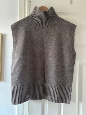 Beige stickad väst från Wera - Ny utan prislapp!! Stickad väst från Wera i beige nyans med hög, ribbad polokrage och bred ribbad kant nedtill. Modellen är ärmlös och har en rak, loose passform.